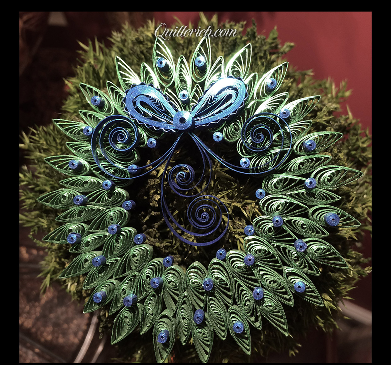&nbsp;Quilled wreath shape with bow and ribbon, green and blue design full view against pine wreath backdrop.